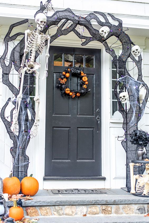 a Halloween porch with black spiderwebs, skeletons and skulls, a black and orange pumpkin wreath on the door