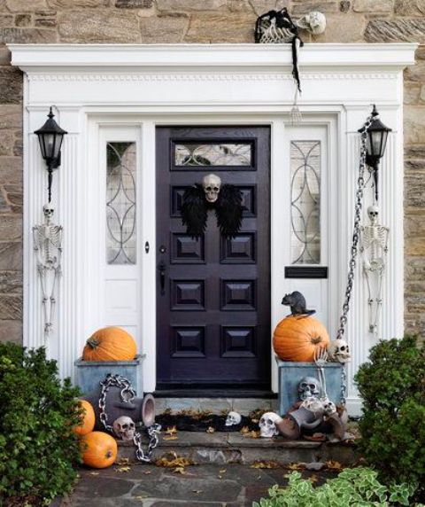 a Halloween porch with natural pumpkins, chains and skulls, a skull and black wings on the door instead of wreath