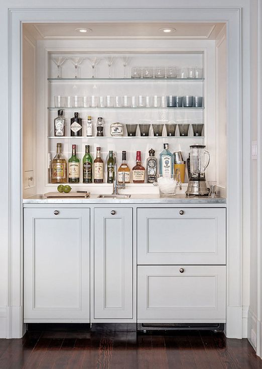a beautiful dove grey home bar with open glass shelves, shaker style cabinets, stone countertops, lots of wine bottles and wine glasses
