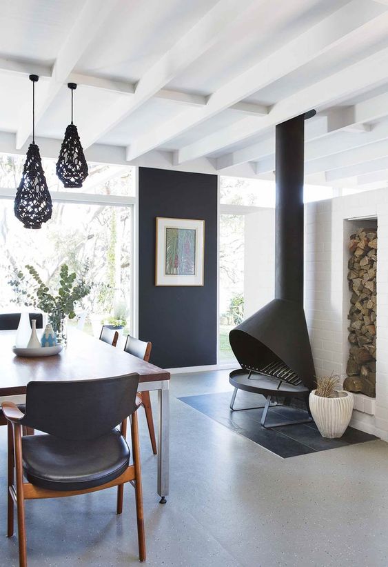 a beautiful modernist dining room with a black accent wall, a black Malm fireplace, black chairs, a stained dining table and black pendant lamps