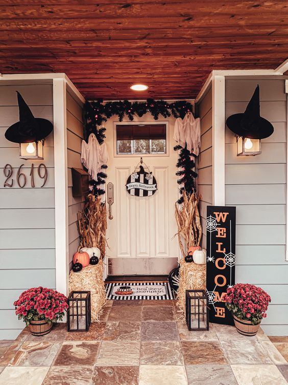 a bold Halloween porch with hay, orange, white and black pumpkins, bold blooms, corn husks, witches' hats and evergreens