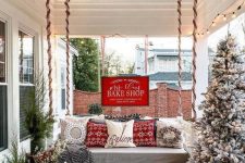 a bright rustic Christmas porch with a flocked Christmas tree with lights, mini trees in buckets, red pillows and a wooden table