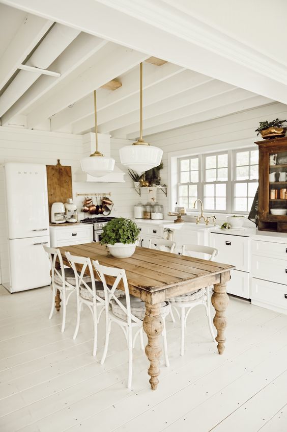 a charming white cottage kitchen with shaker style cabinets, a dining space with a stained table and vintage chairs plus pendant lamps