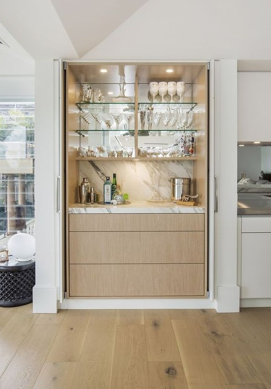 a chic built-in home bar with a marble backsplash, a mirror, glass shelves and sleek drawers for a modern space