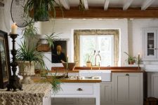 a cottage kitchen with wooden beams, white shaker cabinets and butcherblock countertops, some art and lots of potted plants