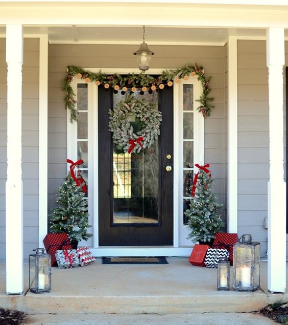 a cozy Christmas front porch with an evergreen and pinecone garland, a snowy wreath, mini trees with lights and lots of gift boxes