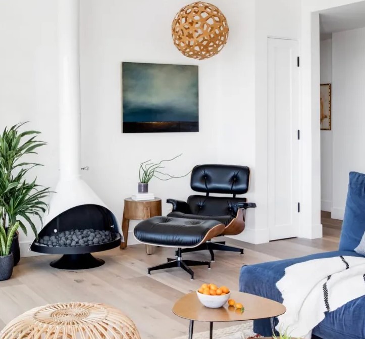 a modern living room with a blue low sofa, a black leather chair with a footrest, a white Malm fireplace and a wooden pendant lamp plus a pouf