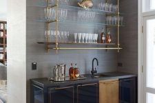 a pretty art deco home bar with a beautiful gold and glass wall mounted shelf, shiny navy cabinetry, a sink with a black faucet