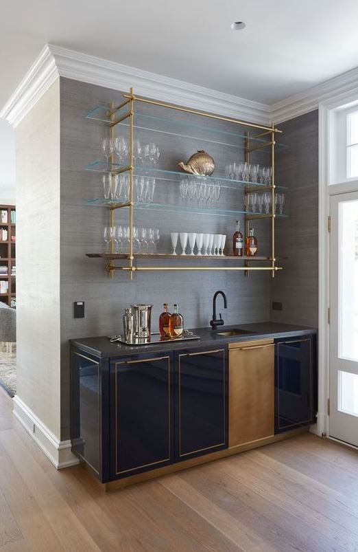 a pretty art deco home bar with a beautiful gold and glass wall mounted shelf, shiny navy cabinetry, a sink with a black faucet