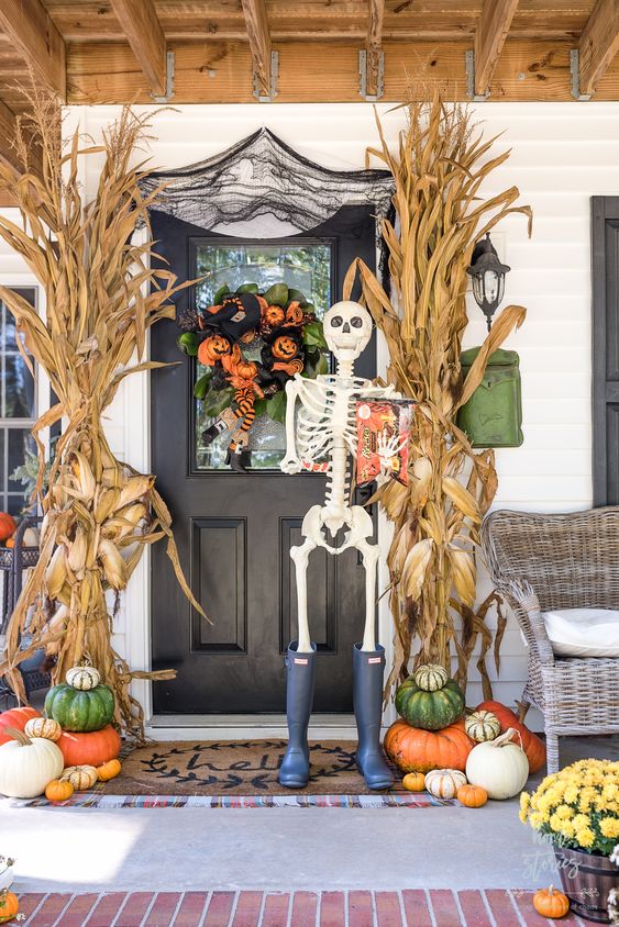 a rustic Halloween porch done with stacked pumpkins, corn husks, a catchy green, orange and black wreath and a skeleton in rubber boots