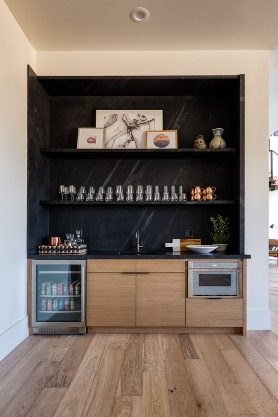 a sleek minimalist home bar with black marble, with open shelves, stained cabinets, a fridge and artwork and a sink with a faucet