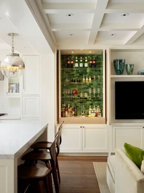 a small built-in home bar with green tiles, glass shelves, built-in lights, a cabinet and a sink plus lots of glasses and wine bottles