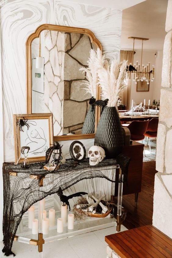 a stylish Halloween console with pillar candles, black cheesecloth, signs and artworks, skulls and blackbirds, a black vase with pampas grass