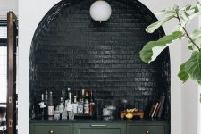 a stylish modern home bar built into a niche, with green cabinetry, a black stone countertop, black glazed brick, a pendant lamp and wine bottles
