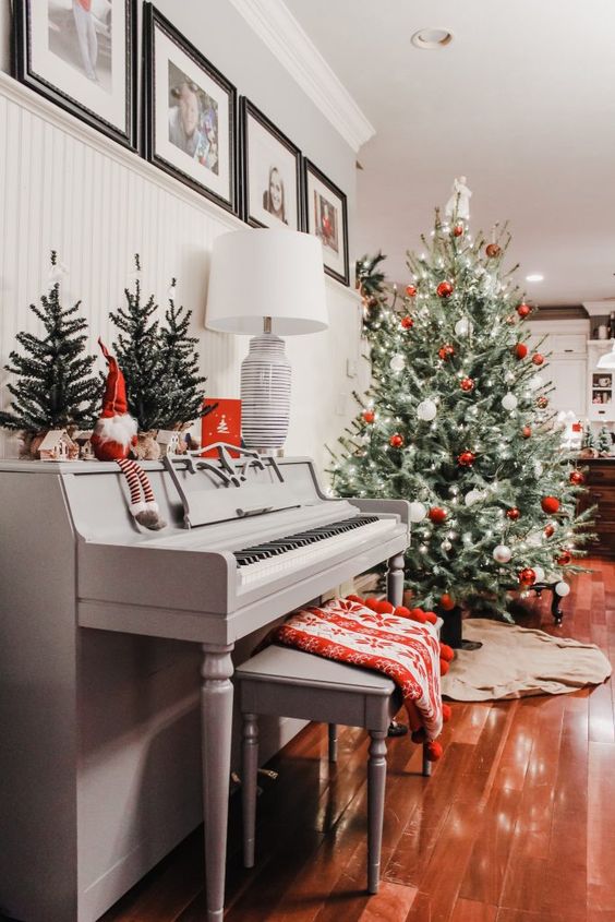a vintage grey piano with some Christmas trees, a gnome, a grey bench with a printed red blanket, a Christmas tree with red and white ornaments