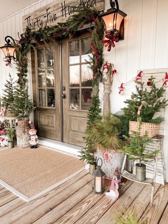 a vintage rustic front porch with mini trees and fir branches in baskets and buckets, candle lanterns, a fir garland, red touches