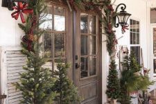 a vintage rustic porch with lots of Christmas trees in baskets, fir branches in a a bucket, lanterns, a fir garland with berries around the door