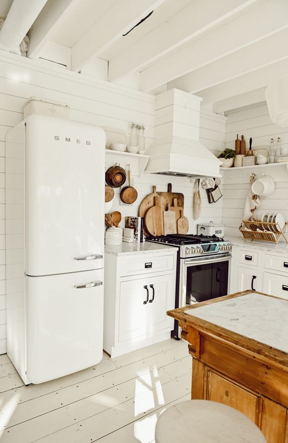 a white cottage kitchen with planked walls and a hood, white shaker cabinets, cool appliances, a stained kitchen island and lots of wood items for decor and usage