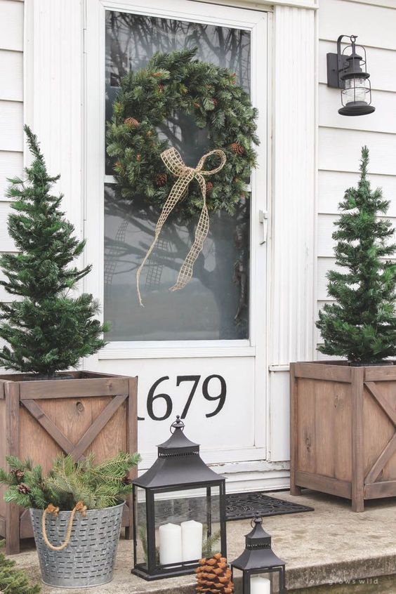 an evergreen wreath with pinecones, Christmas trees, candle lanterns and an evergreen arrangement in a bucket