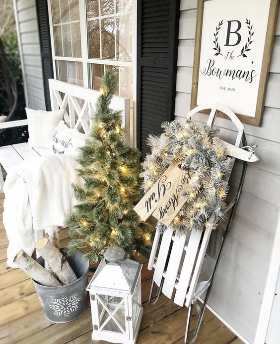 elegant white farmhouse Christmas decor with a white sleigh with a flocked wreath and lights, a white lantern, a mini tree with lights, a bucket with branches and a white bench with a blanket