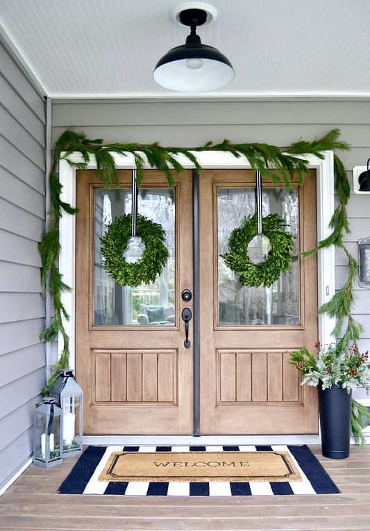 simple natural Christmas front door styling with an evergreen garland and wreaths, candle lanterns and a pale greenery arrangement