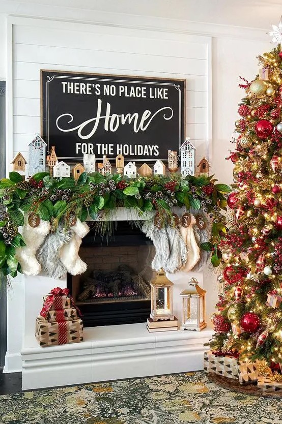 a Christmas mantel with an evergreen and snowy pinecone garland, mini houses and stockings plus a Christmas sign with matching decor