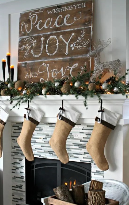 a beautiful rustic Christmas mantel styled with tine balls, silver ornaments, pinecones, evergreens, burlap stockings and wooden plaque Christmas sign