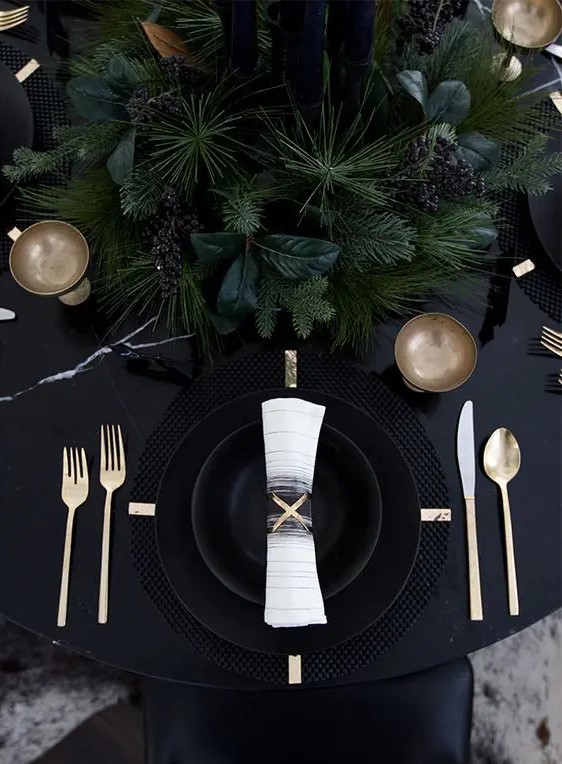 a black table with black plates and chargers, lush evergreens and gold cutlery for a moody NYE tablescape