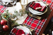 a bold Christmas table setting with a red plaid tablecloth and napkins, woven chargers, white porcelain, fir branches, berries, pomegranates and pinecones