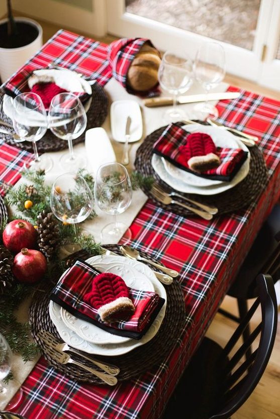 a bold Christmas table setting with a red plaid tablecloth and napkins, woven chargers, white porcelain, fir branches, berries, pomegranates and pinecones