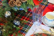a bright holiday table setting with a plaid runner and chargers, pinecones, evergreens, printed ornaments and blue porcelain and glasses
