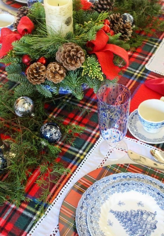 a bright holiday table setting with a plaid runner and chargers, pinecones, evergreens, printed ornaments and blue porcelain and glasses