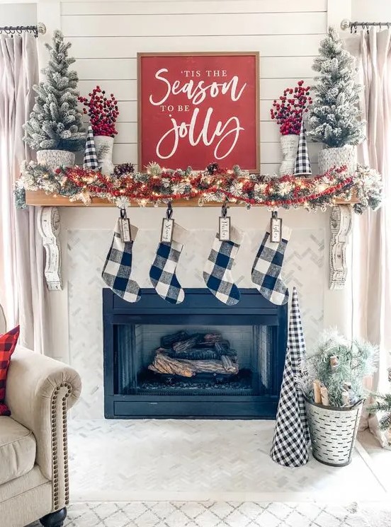 a cozy farmhouse christmas mantel with a snowy pinecone and light garland, red berries, snowy mini trees and buffalo check stockings