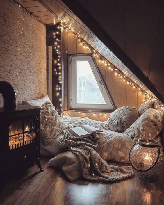 a cpzy attic sleeping space with a bed on the floor, lots of pillows, string lights all over the space and a hearth with lights