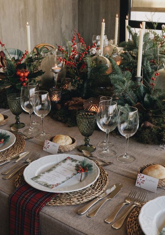a fabulous rustic Christmas tablescape with lush evergreens, berries, candles, green glasses, woden placemats, red plaid napkins