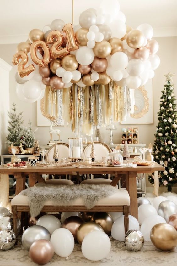 a gold and white NYE party tablescape with cone trees, candles, a balloon arrangement with numbers and Christmas trees around