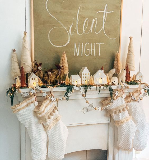 a holiday mantel with white tasseled stockings, an evergreen and light garland, stars and wooden beads, small houses and Christmas trees