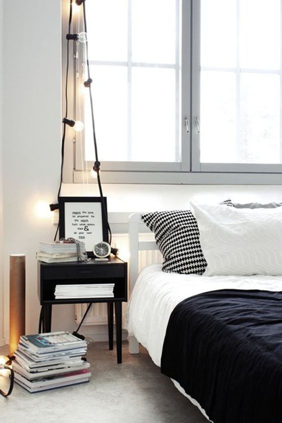 a laconic Scandinavian bedroom with a white bed, navy and black bedding, a black nightstand, string lights and stacks of magazines