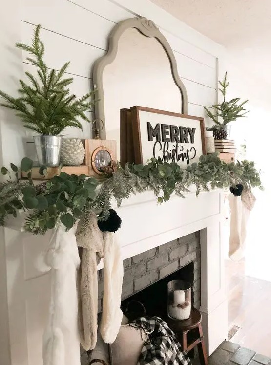 a lovely Christmas mantel with an evergreen and greenery garland, mini Christmas trees, a mirror, a sign and some pinecones is a chic idea