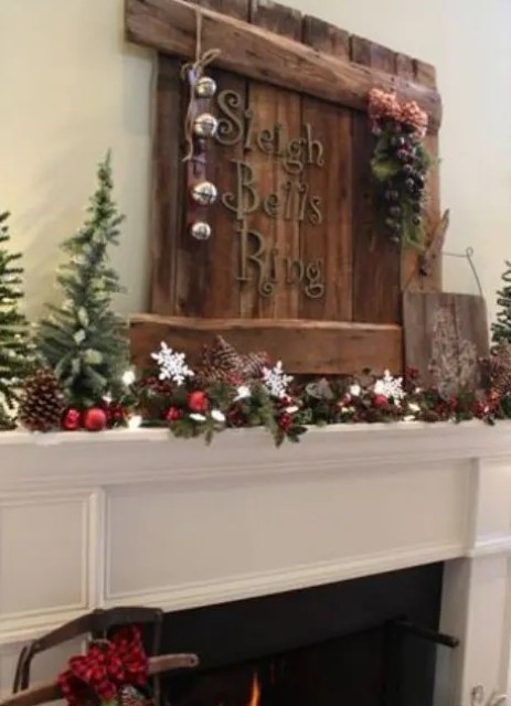 a lovely rustic Christmas mantel with a wooden sign with bells, an evergreen garland with pinecones, berries, ornaments and mini Christmas trees