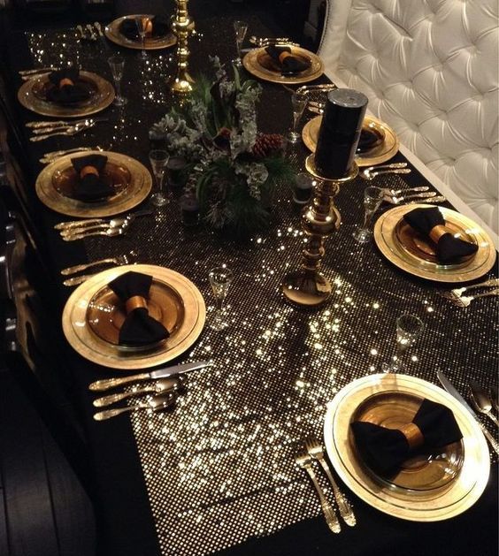 a luxurious glam Christmas tablescape with a shiny tablecloth, gold chargers and cutlery, black napkins and candles and a fir and pinecone centerpiece