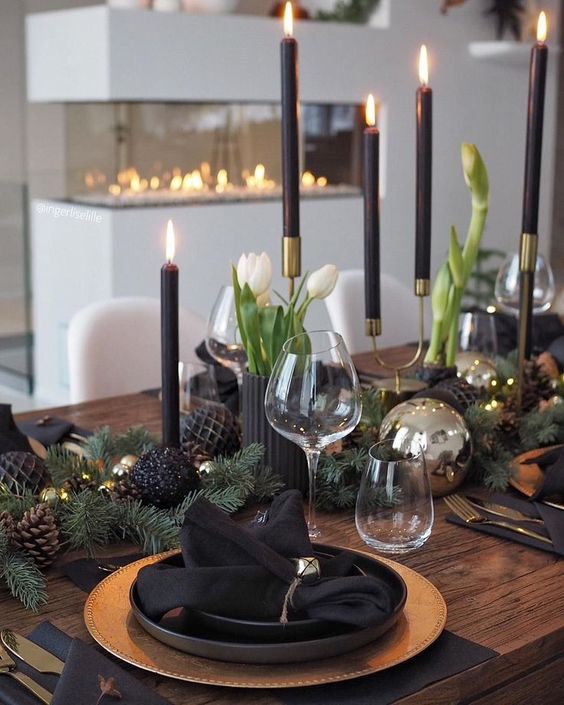 a modern holiday table with gold charger, cutlery, black and shiny ornaments, black candles and white blooms plus bells