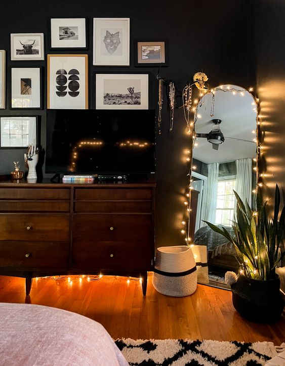 a moody bedroom with black walls, a dark stained dresser, a gallery wall, a curved mirror and string lights here and there