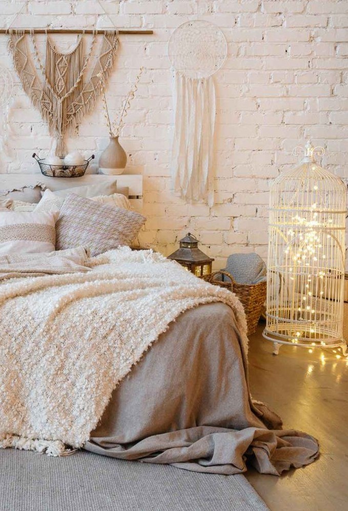 a neutral boho bedroom with a white planked bed and neutral bedding, a cage with lights inside as a light source, some fringe and boho hangings