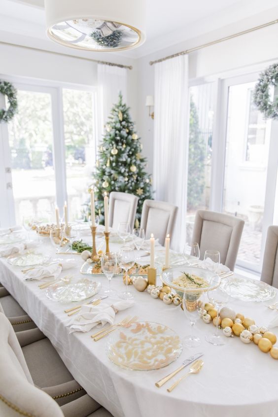 a pretty and bright holiday table setting with gold and white chargers, gold cutlery, white and gold ornaments, gold candleholders and white candles