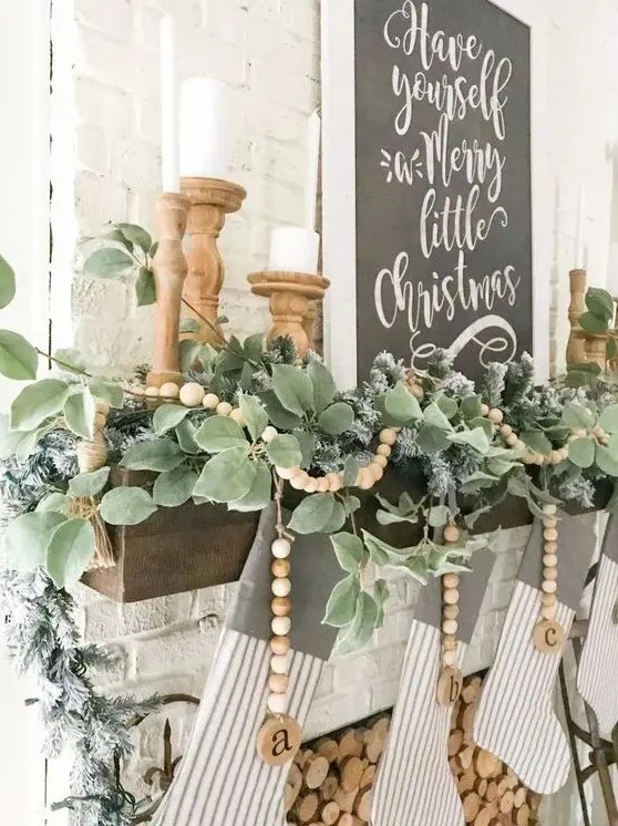 a pretty farmhouse Christmas mantel with a greenery garland and wooden beads, candles in wooden candleholders and striped stockings plus a black and white sign