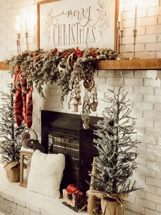 a rustic Christmas mantel wih a snowy evergreen garland with lights, stockings and antlers and mini Christmas trees next to the fireplace