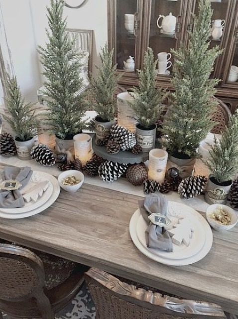 a rustic Christmas table setting with usual and snowy pinecones, potted Christmas trees, wooden Christmas trees and grey napkins