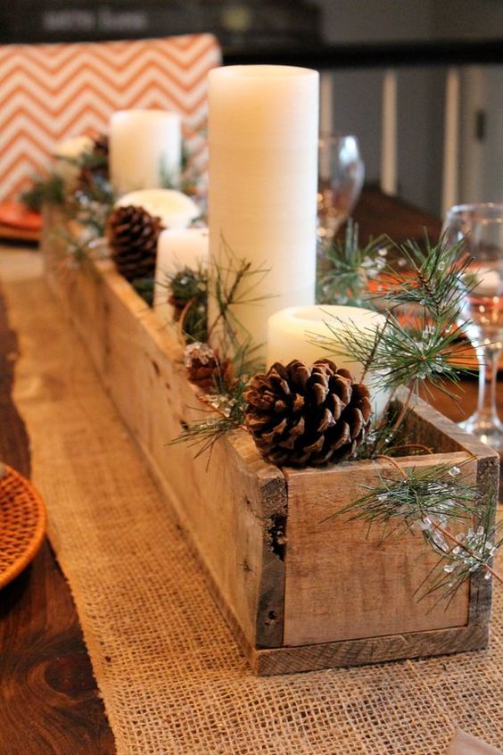 a rustic burlap runner, a wooden planter with evergreens, pinecones and pillar candles as a rustic Christmas centerpiece