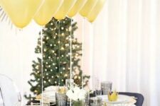 a shiny black and gold NYE party tablescape with a black tablecloth, gold chargers and gold balloons over the table, grey glasses and white blooms